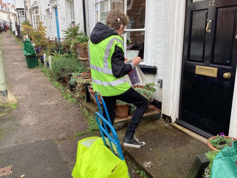 A student wearing a high visibility jacket is folding up a copy of the Exmouth Journal, ready to deliver it through the letterbox on a front door.